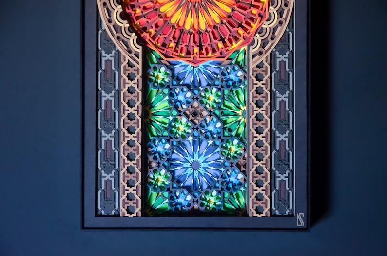 May include: A framed paper art piece with a colourful geometric design. The design features a central circle with radiating lines, surrounded by intricate patterns in shades of red, orange, yellow, green, blue, and brown.