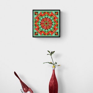 May include: A framed geometric art print with a red, green, and orange pattern. The print is hanging on a white wall above a red vase with a green plant and a red bird figurine.