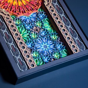 May include: A framed artwork featuring intricate, layered paper cutouts in shades of blue, green, orange, and red. The design incorporates geometric patterns and floral motifs.