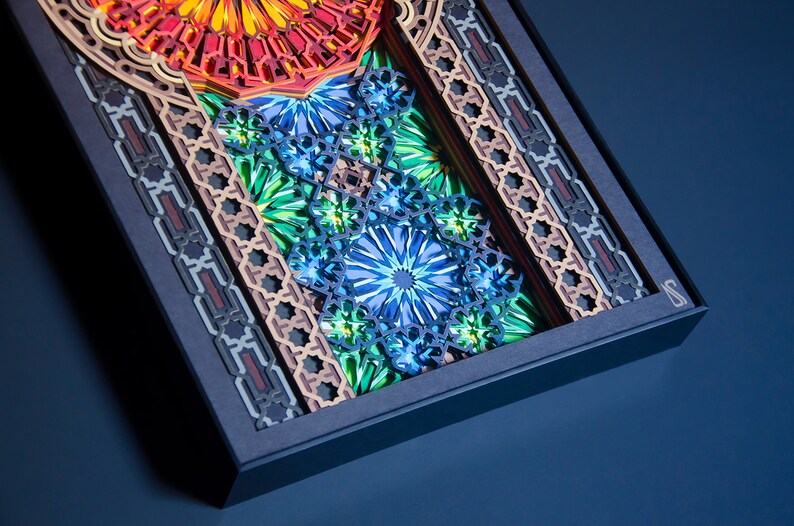 May include: A framed artwork featuring intricate, layered paper cutouts in shades of blue, green, orange, and red. The design incorporates geometric patterns and floral motifs.