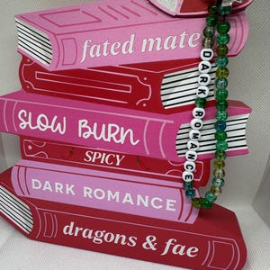 May include: A stack of decorative wooden books in shades of pink and red, with titles like "Fated Mate" and "Dark Romance." A beaded charm with the words "DARK ROMANCE" hangs from the top. A glittery heart shape is attached to the top.