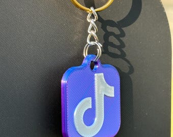 May include: A purple and blue keychain featuring the TikTok logo in white. The keychain has a silver chain and a gold-colored ring. The background is a dark, solid color.