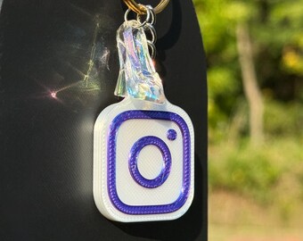 May include: A white and purple Instagram logo keychain with a gold keyring. The keychain has a clear, iridescent top. The Instagram logo is a white square with a purple border and a purple circle in the center.