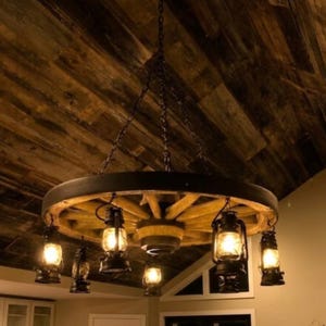 May include: A rustic wagon wheel chandelier with six hanging lanterns. The wheel is made of wood and has a dark metal rim. The lanterns are black and have glass panels, emitting a warm, inviting glow. The fixture hangs from chains.