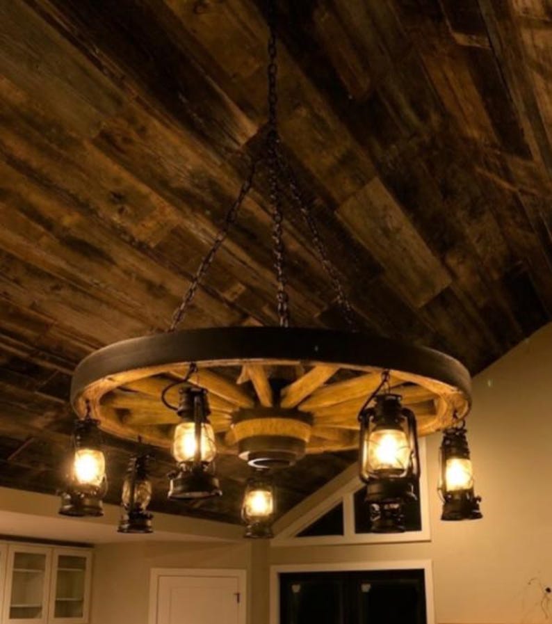 May include: A rustic wagon wheel chandelier with six hanging lanterns. The wheel is made of wood and has a dark metal rim. The lanterns are black and have glass panels, emitting a warm, inviting glow. The fixture hangs from chains.