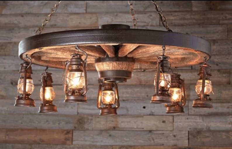 May include: Rustic wagon wheel chandelier with eight hanging oil lanterns. The lanterns have clear glass and are lit with warm, inviting light. The wheel is made of wood and metal, creating a vintage aesthetic.
