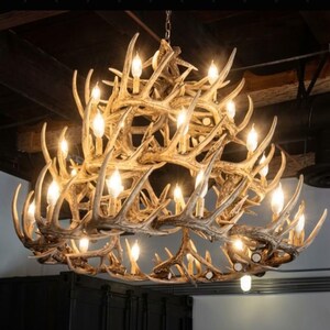 May include: A large, rustic chandelier crafted from multiple deer antlers. The chandelier has multiple light bulbs, creating a warm glow. The antlers are a natural brown color, and the fixture hangs from a chain.