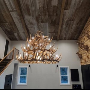 May include: A large antler chandelier hangs from a wood-paneled ceiling. The chandelier is made of multiple deer antlers and has several light bulbs. The room has a staircase and windows.
