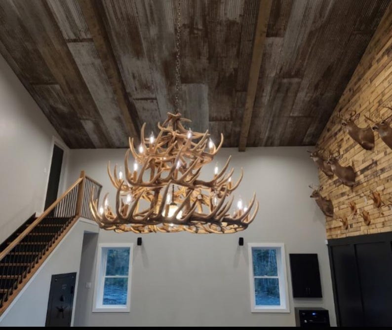 May include: A large antler chandelier hangs from a wood-paneled ceiling. The chandelier is made of multiple deer antlers and has several light bulbs. The room has a staircase and windows.