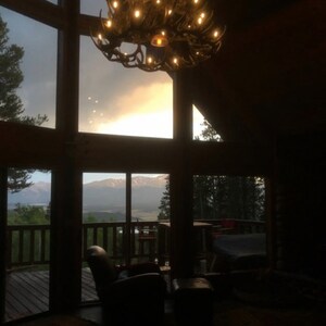May include: A rustic interior with a large window framing a mountain view at sunset. A chandelier made of antlers hangs from the ceiling. The room features a leather armchair and a wooden deck.