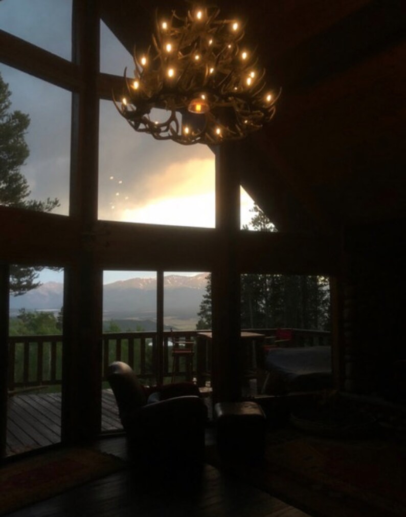 May include: A rustic interior with a large window framing a mountain view at sunset. A chandelier made of antlers hangs from the ceiling. The room features a leather armchair and a wooden deck.