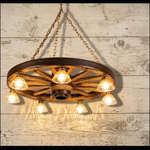 Puede incluir: Una l&aacute;mpara de ara&ntilde;a r&uacute;stica con forma de rueda de carro con siete luces. La rueda de madera est&aacute; te&ntilde;ida de marr&oacute;n y las luces tienen pantallas de cristal color &aacute;mbar. La l&aacute;mpara cuelga de una cadena contra una pared de madera.