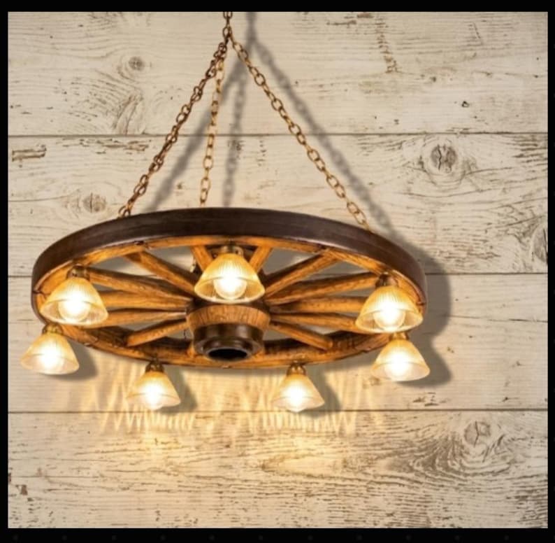 Puede incluir: Una l&aacute;mpara de ara&ntilde;a r&uacute;stica con forma de rueda de carro con siete luces. La rueda de madera est&aacute; te&ntilde;ida de marr&oacute;n y las luces tienen pantallas de cristal color &aacute;mbar. La l&aacute;mpara cuelga de una cadena contra una pared de madera.