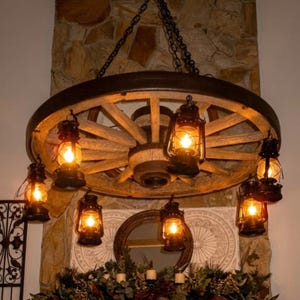 May include: A rustic wagon wheel chandelier with six oil lanterns. The wheel is made of wood and has a dark metal rim. The lanterns are black and have a warm, inviting glow. The chandelier hangs from a chain.