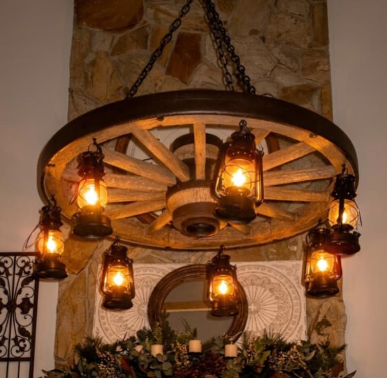 May include: A rustic wagon wheel chandelier with six oil lanterns. The wheel is made of wood and has a dark metal rim. The lanterns are black and have a warm, inviting glow. The chandelier hangs from a chain.