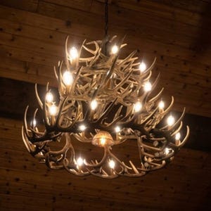 May include: A rustic antler chandelier with multiple light bulbs. The chandelier is made of brown antlers and hangs from a chain. The light bulbs are lit, casting a warm glow. The background is a wooden ceiling.