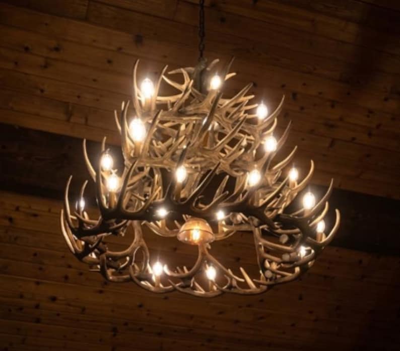 May include: A rustic antler chandelier with multiple light bulbs. The chandelier is made of brown antlers and hangs from a chain. The light bulbs are lit, casting a warm glow. The background is a wooden ceiling.