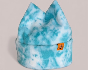May include: A blue and white tie-dye beanie hat with a folded brim. The hat has two pointed peaks at the top and a small orange tag with a graphic. The hat is made of a soft, fuzzy material.