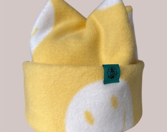 May include: A yellow and white fleece hat with cat ears. The hat features a white design on the front and ears, with a small green tag on the side. The hat is designed to be worn on the head.