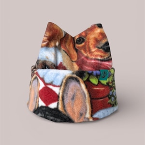 May include: A fleece hat featuring a detailed print of a dachshund dog. The hat has a folded brim and a top that resembles the dog's ears. The design includes a red and white pattern, a flower pot, and a blue and green floral arrangement.