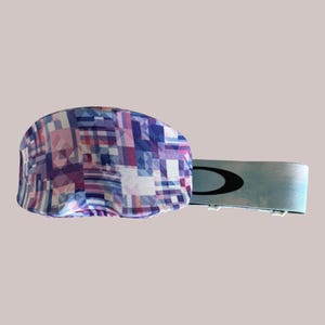 May include: A patterned eye mask with a geometric design in shades of pink, purple, and blue. The mask has a light blue strap with a black logo. The mask is designed to block out light.