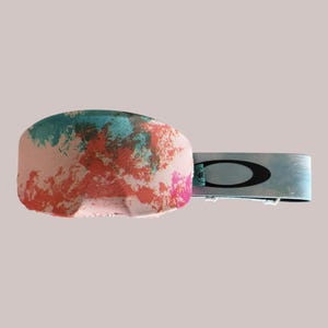 May include: A pair of Oakley goggles with a colorful, abstract design. The goggles feature a pink base with splashes of teal, orange, and pink. The strap is light blue with the Oakley logo in black.