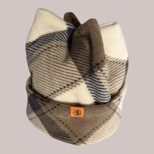 May include: A plaid fleece beanie in cream, brown, and navy blue. The hat features a folded brim and a small, square brown patch with a logo. The beanie is designed to keep the head warm.