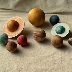May include: A collection of hand-painted wooden planets, including Earth, Jupiter, and Mars, arranged on a neutral-toned surface. The planets vary in size and color, with detailed surface patterns. A large, yellow sun is also present, creating a miniature solar system display.