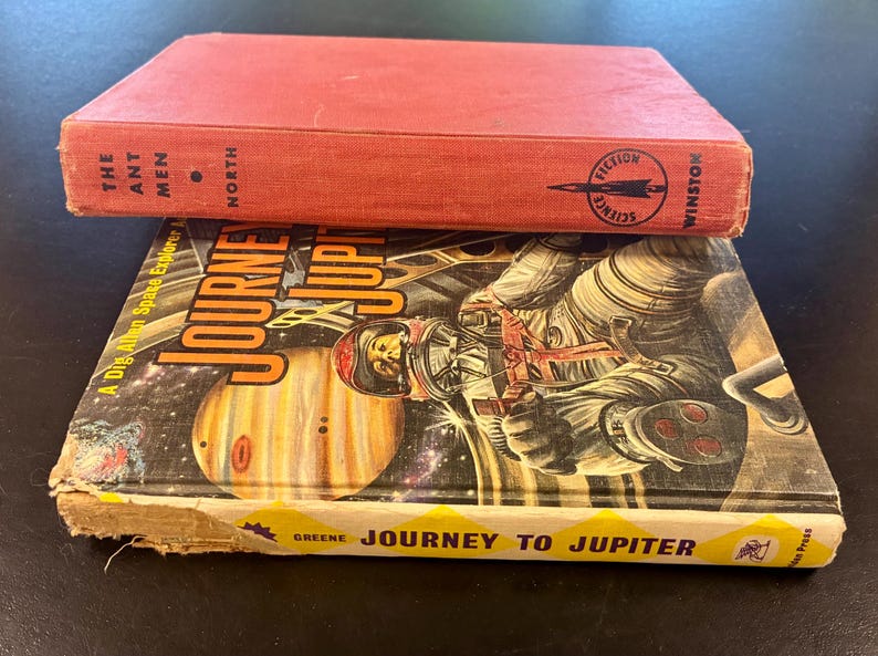 Op de afbeelding: Twee vintage sciencefictionboeken. Het bovenste boek is rood met de titel "The Ant Men" en de uitgever "Winston". Het onderste boek heet "Journey to Jupiter" met een illustratie van een astronaut en de planeet Jupiter.