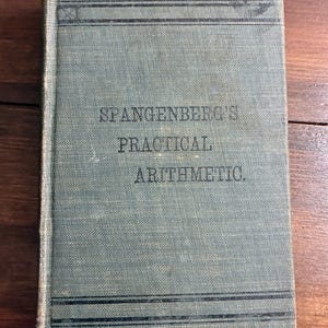 May include: A vintage hardcover book titled Spangenberg's Practical Arithmetic with a faded blue-grey cloth cover. The front cover features dark horizontal lines at the top and bottom, with the title printed in a simple, dark serif font.