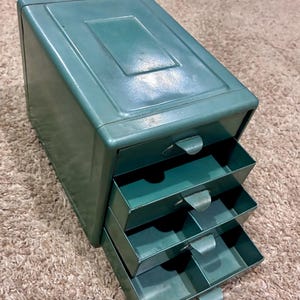May include: A teal metal storage unit featuring four stacked drawers, each equipped with a small protruding tab handle. The rectangular cabinet has a smooth, solid-colored finish and a flat top surface, providing a compact organization solution.
