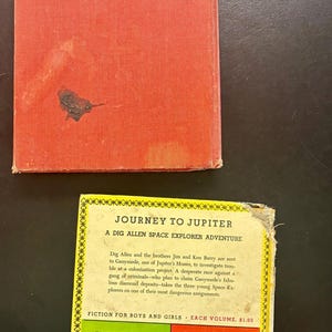 Op de afbeelding: Vintage kinderboek "Reis naar Jupiter" met een rode kaft en een kleurrijk ge&iuml;llustreerde achterkant. Het boek bevat avonturenverhalen voor jongens en meisjes, waaronder "Dig Allen Space Explorer Adventures".