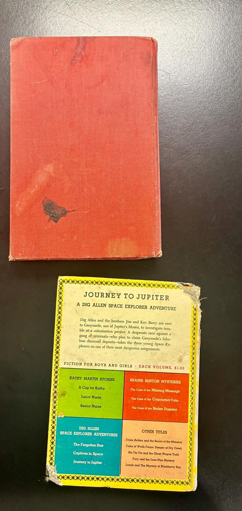 Op de afbeelding: Vintage kinderboek "Reis naar Jupiter" met een rode kaft en een kleurrijk ge&iuml;llustreerde achterkant. Het boek bevat avonturenverhalen voor jongens en meisjes, waaronder "Dig Allen Space Explorer Adventures".