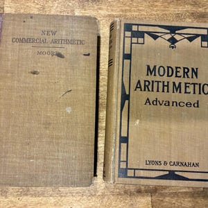 May include: Two vintage hardcover textbooks with tan cloth covers. The left book is titled New Commercial Arithmetic by Moore with a dark red spine. The right book is titled Modern Arithmetic Advanced by Lyons & Carnahan with black border art.