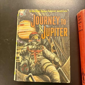 Op de afbeelding: Vintage boekomslag getiteld "Journey to Jupiter" met een astronaut in een ruimtepak, met de planeet Jupiter op de achtergrond. De omslag heeft een retro-ontwerp met oranje en gele letters en illustraties.