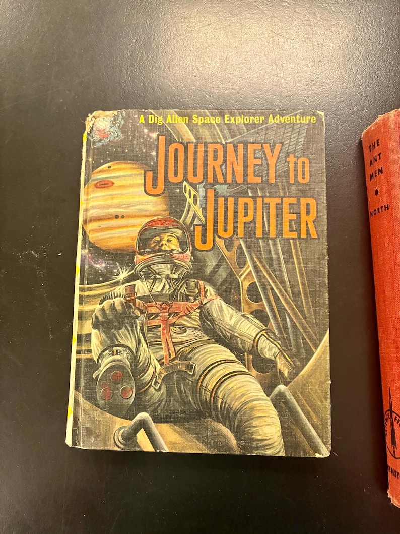 Op de afbeelding: Vintage boekomslag getiteld "Journey to Jupiter" met een astronaut in een ruimtepak, met de planeet Jupiter op de achtergrond. De omslag heeft een retro-ontwerp met oranje en gele letters en illustraties.