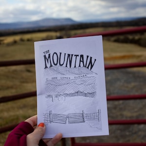 Può includere: Una copertina di fumetto in bianco e nero con il titolo "The Mountain" di Rose Comics Turner. La copertina presenta un disegno di una catena montuosa con una recinzione e una piccola casa in primo piano.