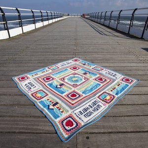 May include: A colourful crocheted blanket with a beach theme. The blanket is laid out on a wooden pier overlooking the sea. The blanket features a variety of designs, including seashells, lighthouses, and waves. The text "Wish you were here" and "With love" is written on the blanket.
