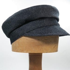 May include: A dark denim-colored cap with a structured design, featuring a flat top and a short, angled brim. The hat is displayed on a wooden hat form against a white background. This classic style is suitable for various outfits.