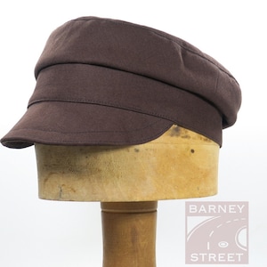 May include: A solid dark brown newsboy cap featuring a structured crown, a wide horizontal band, and a short, curved brim. The cap is displayed on a wooden form, and a logo with the text BARNEY STREET is visible in the lower right corner.