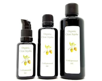 May include: Three bottles of organic clear jojoba oil, cold pressed. The bottles are black with white labels and a drawing of a jojoba plant. The bottles are arranged in order of size, with the largest bottle on the right and the smallest bottle on the left.