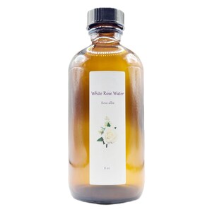 May include: A brown glass bottle of White Rose Water, with a black cap. The label features the text "White Rose Water" and "Rosa alba", along with a white rose illustration. The bottle contains 8 oz of product.