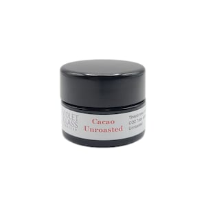 May include: A small, round, black jar with a black lid. The jar has a white label with the words "VIOLET GLASS FOREVER" and "Cacao Unroasted" in red text. The jar contains a dark substance.