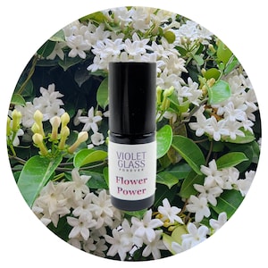 Puede incluir: Un frasco negro de rodillo con una etiqueta blanca que dice "Violet Glass Forever Flower Power". El frasco está delante de un fondo de flores blancas.