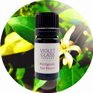 May include: A small, dark brown glass bottle with a black cap. The bottle is labelled "VIOLET GLASS FOREVER Petitgrain Sur Fleurs". The bottle is sitting on a green and yellow background with a white flower.