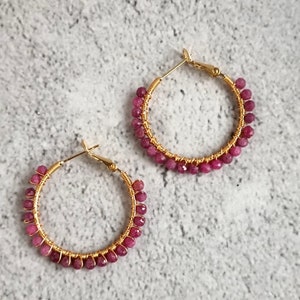 Peut inclure: Une paire de créoles dorées ornées de petites perles de pierres précieuses facettées rose foncé enroulées autour de la structure en fil métallique. Chaque boucle d'oreille possède un fermoir à charnière sur le dessus, les perles entourant tout le cerceau.