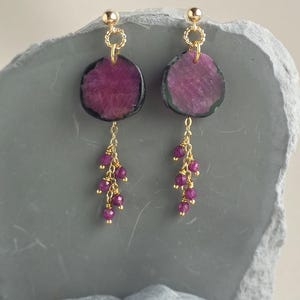 May include: A pair of gold-tone earrings featuring circular, dark-rimmed magenta stone slices suspended from gold posts. Each earring includes a delicate gold chain tassel adorned with small, faceted magenta beads hanging from the central stone.