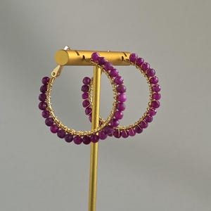 May include: A pair of hoop earrings featuring small, faceted, deep magenta-colored beads wrapped around a thin gold-toned wire frame. The circular earrings are displayed on a gold-toned T-shaped stand, showcasing the vibrant purple-pink gemstone beads.