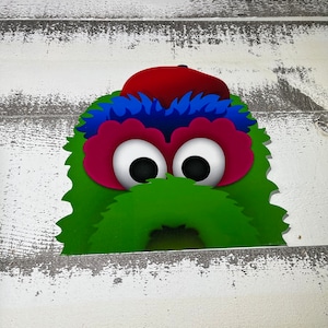 May include: A green, fuzzy monster with a red hat, blue hair, and large eyes peeking out from behind a white wooden surface.