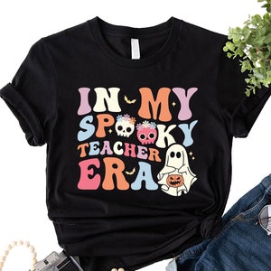 Puede incluir: Camiseta negra con un gráfico colorido que dice "In My Spooky Teacher Era" con un fantasma y calaveras.